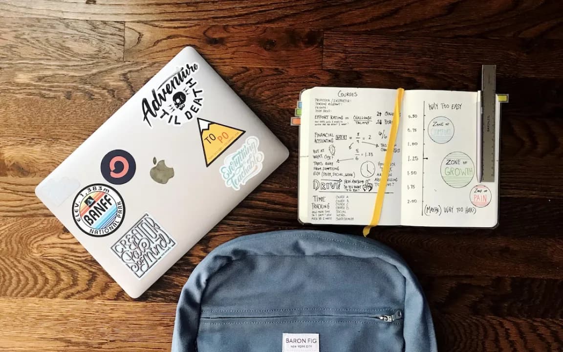 A laptop, an open notebook of hand-drawn study charts and a backpack laid out on a wooden floor.