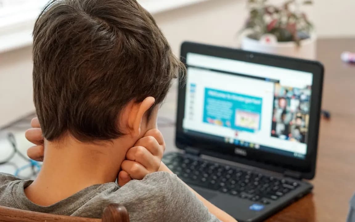 A boy seen from behind at a laptop, following an online class with a grid of other pupils on screen.