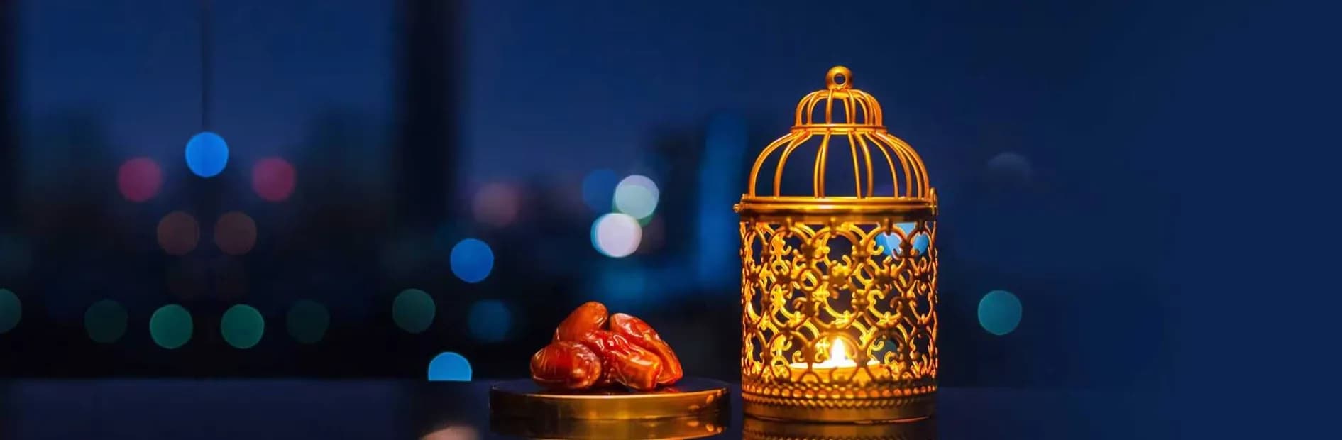 A lit brass lantern beside a plate of dates on a windowsill at dusk, with city lights blurred behind.