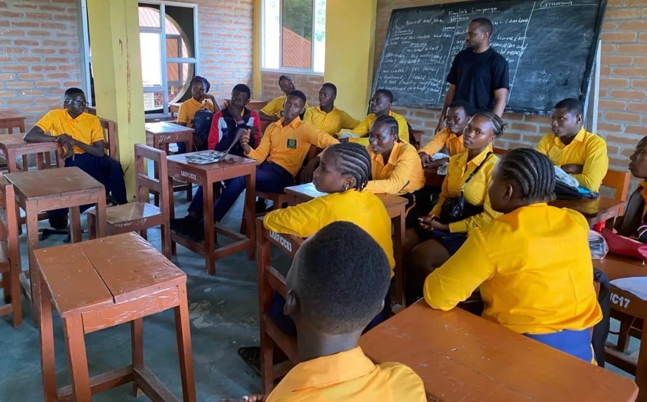 Students in yellow uniforms seated at wooden desks in a classroom at Lamin and Kadie Bah Academy, facing a blackboard of English grammar notes where a member of the Muallemi team is teaching.
