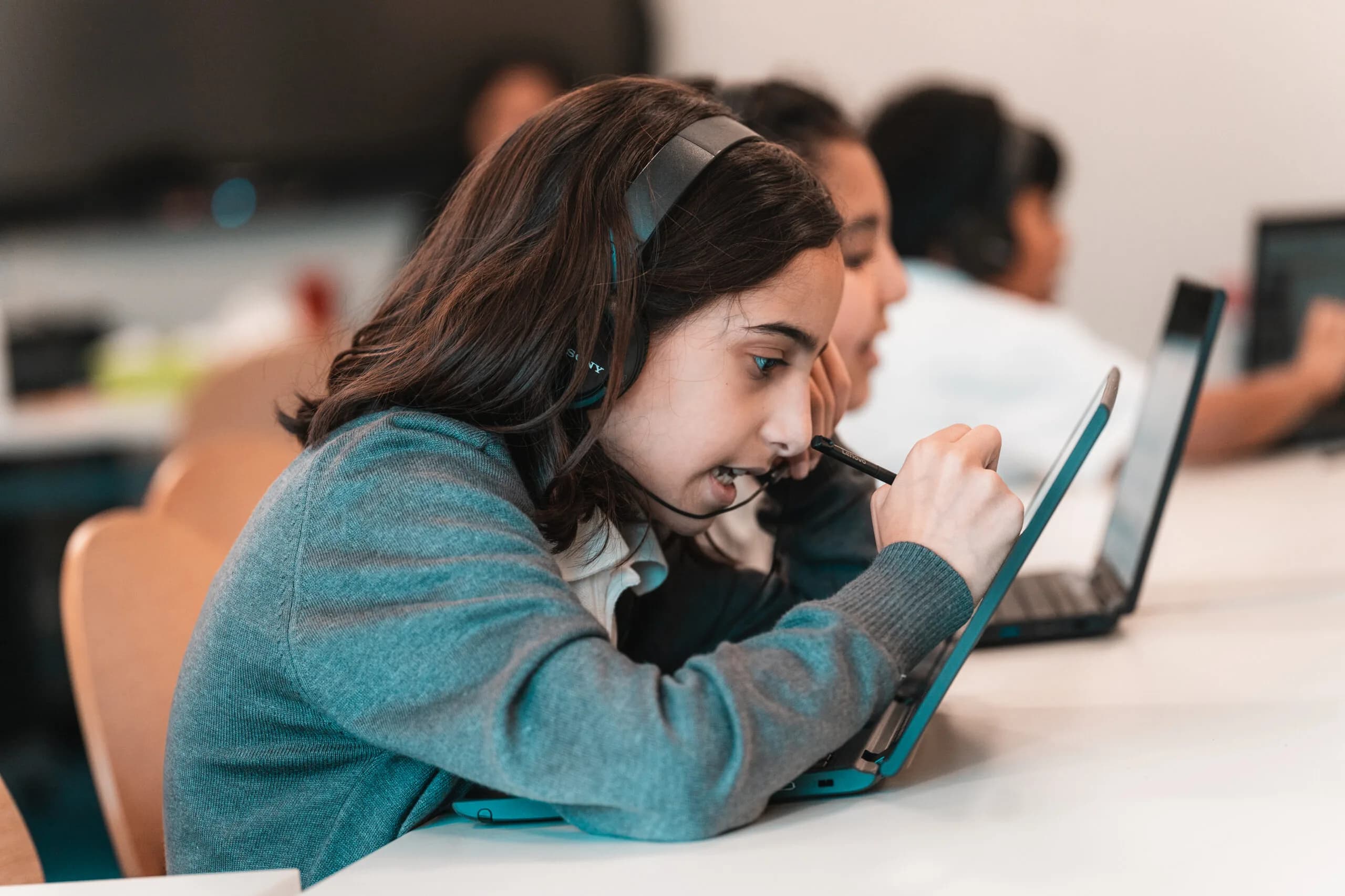 A girl wearing a headset writes on a laptop screen with a stylus during an online lesson, with other learners at laptops behind her.